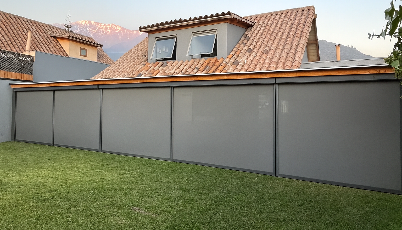 Cierre de terraza Ziptrak con tela screen completamente desplegado, estructura de aluminio oscuro, protegiendo un comedor exterior protegido, rodeado de césped.