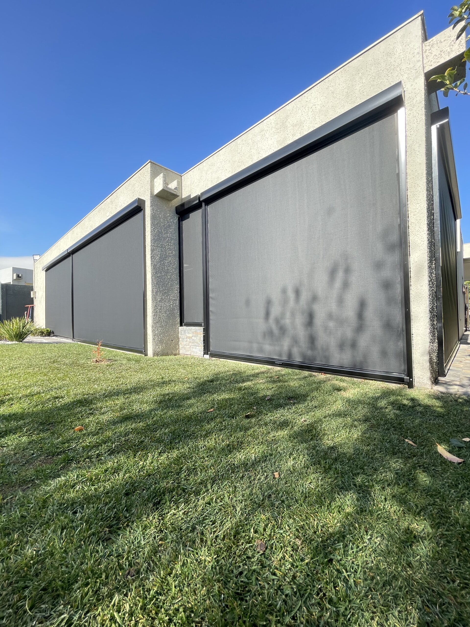 Cierre de terraza Ziptrak con tela screen completamente desplegado, estructura de aluminio oscuro, protegiendo un comedor exterior protegido, rodeado de césped.